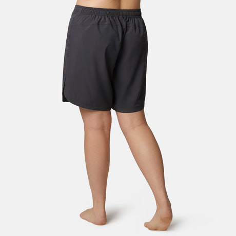 A person stands barefoot on a white background, shown from the back, wearing Free Country Women's Plus Size Bermuda Board Short II in dark gray with an elastic waistband and quick-drying fabric.