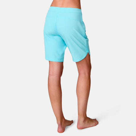 A woman stands barefoot on a white background, shown from the back, wearing light blue Women's Bermuda Board Short II.