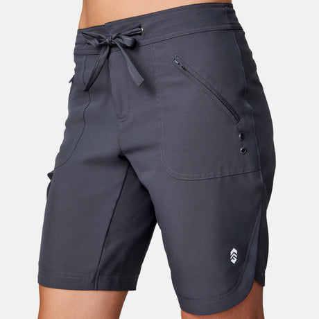 A person is wearing Women's Bermuda Board Short II in dark gray, featuring a front drawstring, two zippered side pockets, a small white logo by the left hem, and quick-drying fabric for comfort.