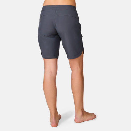 A woman stands barefoot, facing away, wearing Women's Bermuda Board Short II in dark gray with UPF 50+ protection against a plain white background.