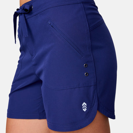 [Model is wearing a size Small.] Woman wearing Free Country Women's 5" Bermuda Board Short in Marine, size S, made from quick drying UPF stretch performance fabric featuring a fly front zipper closure with a drawcord and two front pockets 