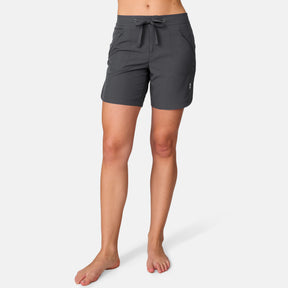 A woman models the Free Country Women's 7" Bermuda Board Short in dark gray, featuring front pockets, a drawstring waist, and a quick-dry fabric with UPF 50+ protection. She stands barefoot against a plain white background.