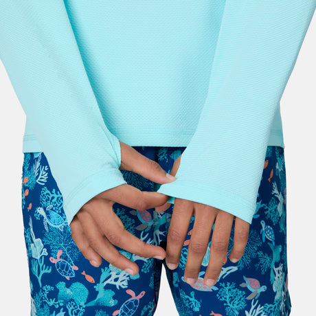 A person wears the Free Country Kids' Everybody SunFree UPF Hoodie in light blue with thumb holes and blue swim shorts made of recycled polyester, featuring a sea life pattern with turtles, fish, and coral. Their hands are clasped in front.