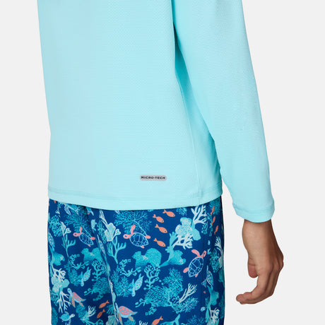 A child wears the Free Country Kids' Everybody SunFree UPF Hoodie in light blue, paired with blue shorts featuring a vibrant sea life pattern; only the torso and one arm are shown.