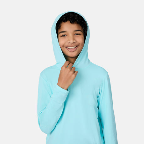 A smiling young person with curly hair pulls up the hood of The Kids' Everybody SunFree UPF Hoodie by Free Country, made from recycled polyester, standing against a plain white background.