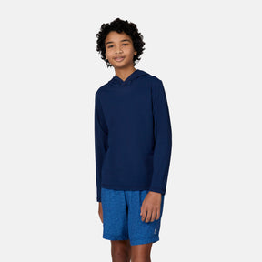 A smiling boy with curly hair wears the Free Country Kids' Everybody SunFree UPF Hoodie in navy blue and blue patterned shorts, standing against a plain light gray background.