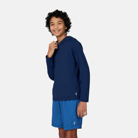 A smiling boy with curly hair wears the Free Country Kids' Everybody SunFree UPF Hoodie in navy, made from recycled polyester, paired with blue shorts, standing against a plain light gray background.