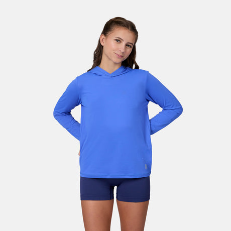 A young person with long brown hair wears the Free Country Kids' Everybody SunFree UPF Hoodie in blue and navy shorts, standing against a plain white background with hands on hips and a slight smile.