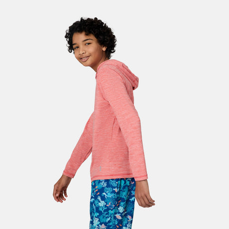A smiling young person with curly hair wears the Free Country Kids' Everybody SunFree UPF Hoodie in pink and blue patterned shorts, standing sideways and looking at the camera against a plain white background, ready for sun protection.