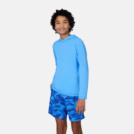 A smiling boy with curly hair wears the Kids' Everybody SunFree UPF Hoodie by Free Country, made from recycled polyester, and blue patterned shorts. He stands against a light background with one hand in his pocket, ready for sun protection.