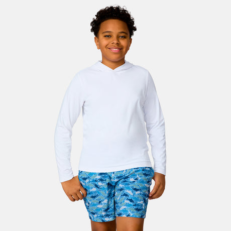 A smiling boy wears The Kids' Everybody SunFree UPF Hoodie by Free Country in white, paired with blue shark-print swim shorts—ideal sun protection for bright days, shown against a plain light background.