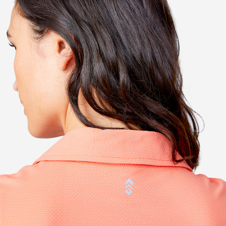 A woman with shoulder-length dark hair is seen from behind wearing the Free Country Women's SunFree Sleeveless Top in coral, made of moisture-wicking fabric with a small white geometric logo below the back collar.