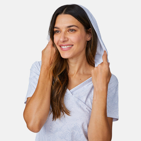 A smiling woman with long brown hair wears the Free Country Women's SunFree UPF Dress, a light-colored, short-sleeve hoodie cover-up made from recycled polyester, holding the hood with both hands against a plain white background.