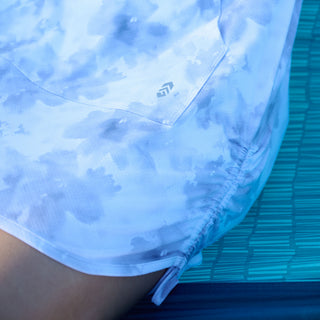 A close-up of a person wearing the Free Country Women's SunFree UPF Dress, featuring a light blue and white floral pattern, a subtle geometric logo, and UPF 50+ sun protection, while sitting on a blue textured surface.