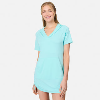 A smiling woman with long blonde hair wears the Free Country Women's SunFree UPF Dress—a light blue, short-sleeve hooded dress with a front pocket, crafted from recycled polyester cover-up fabric and featuring UPF 50+ sun protection—while standing against a plain white background.