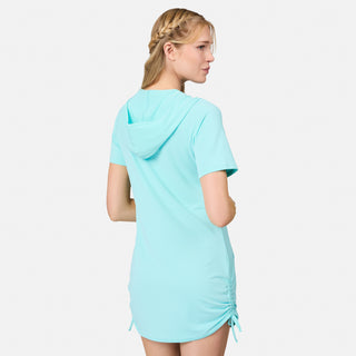 A woman with blonde braided hair is seen from the back against a plain white background, wearing the Free Country Women's SunFree UPF Dress—a light blue, short-sleeve hooded cover-up made from recycled polyester.