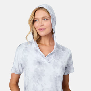 A woman is pictured wearing the Free Country Women's SunFree UPF Dress in light gray, featuring short sleeves, a hood, and a subtle floral pattern. She looks slightly to the side with a smile. Crafted from soft recycled polyester cover-up fabric that provides UPF 50+ sun protection, this dress is ideal for sunny summer days. The background is plain white.