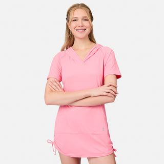 A smiling woman stands with arms crossed, wearing the Free Country Women's SunFree UPF Dress—a short-sleeved, light pink hooded dress with UPF 50+ sun protection and side drawstrings—against a plain white background.