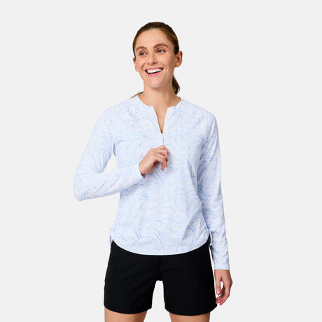 A smiling person stands against a plain background, wearing the Free Country Women's SunFree Quarter Zip UPF Sunshirt in white with a subtle blue pattern and black shorts.