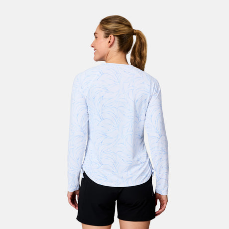 A woman with a ponytail wears the Free Country Women's SunFree Quarter Zip UPF Sunshirt in white and black shorts, standing with her back to the camera and smiling over her shoulder against a plain white background.