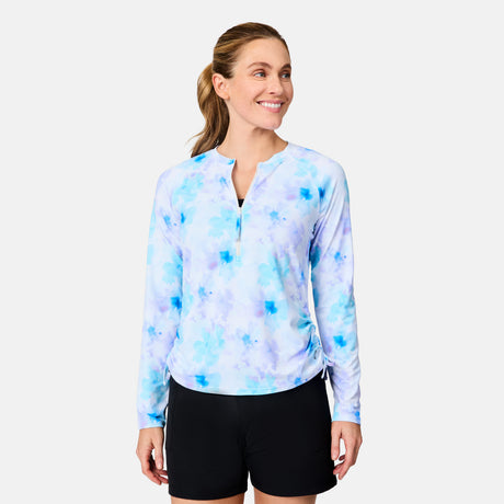 A woman with light brown hair in a ponytail is smiling and looking to the side, wearing the Free Country Women's SunFree Quarter Zip UPF Sunshirt in blue and purple tie-dye with black shorts, against a plain background.