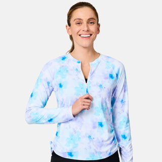 A smiling woman with light brown hair in a ponytail models the Free Country Women's SunFree Quarter Zip UPF Sunshirt, featuring long sleeves and a light blue and purple tie-dye design, unzipping the collar slightly against a white background.