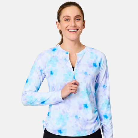 A smiling woman with light brown hair in a ponytail models the Free Country Women's SunFree Quarter Zip UPF Sunshirt, featuring long sleeves and a light blue and purple tie-dye design, unzipping the collar slightly against a white background.