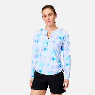 A woman stands against a plain white background wearing the Free Country Women's SunFree Quarter Zip UPF Sunshirt in light blue and white tie-dye with black shorts. The shirt features long sleeves, moisture-wicking fabric, and UPF 50+ sun protection.