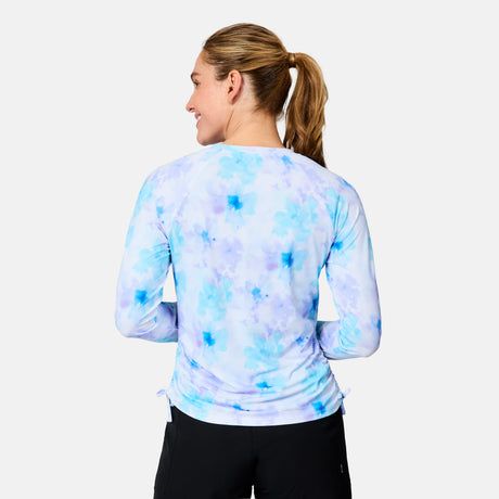 A blonde woman with a ponytail stands with her back to the camera, wearing the Free Country Women's SunFree Quarter Zip UPF Sunshirt in blue-purple tie-dye and black pants, against a plain light background.