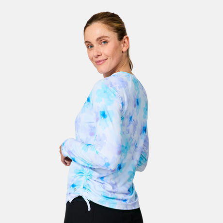 A woman with light brown hair in a ponytail models the Free Country Women's SunFree Quarter Zip UPF Sunshirt, wearing black pants and standing sideways as she looks over her shoulder with a slight smile.