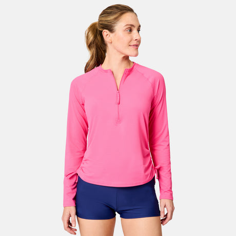 A woman in a bright pink Free Country Women's SunFree Quarter Zip UPF Sunshirt and navy blue shorts stands angled to the side, looking left, against a plain light background.