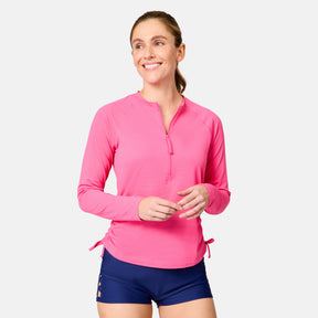 A woman in a bright pink Free Country Women's SunFree Quarter Zip UPF Sunshirt and navy shorts stands smiling with loosely clasped hands, her light brown hair pulled back, against a plain light background.