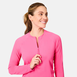 A smiling woman with light skin and brown hair in a ponytail zips up the Free Country Women's SunFree Quarter Zip UPF Sunshirt in bright pink, looking off to her left against a plain light background.