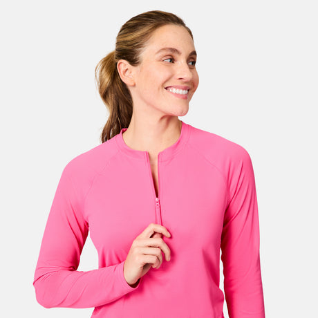 A smiling woman with light skin and brown hair in a ponytail zips up the Free Country Women's SunFree Quarter Zip UPF Sunshirt in bright pink, looking off to her left against a plain light background.