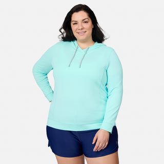 A woman stands smiling with one hand on her hip against a plain white background, wearing the Free Country Women's Plus Size SunFree UPF Hoodie in light blue and navy blue shorts.