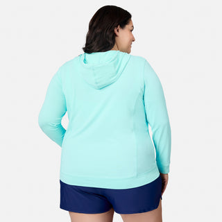 A woman is shown from the back wearing the Free Country Women's Plus Size SunFree UPF Hoodie in light blue, paired with navy shorts against a plain white background. This long-sleeve hoodie features UPF 50+ sun protection and moisture-wicking technology, offering comfort and coverage for active outdoor days.