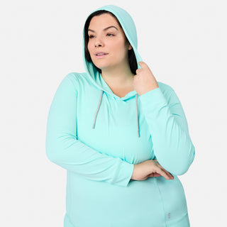 A woman models the Free Country Women's Plus Size SunFree UPF Hoodie in light blue, featuring UPF 50+ sun protection. She stands against a plain white background, gazing to the side with one hand holding her hood and her other arm crossed.