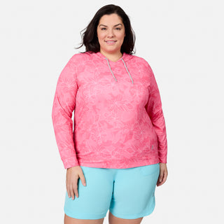 A woman smiles at the camera while wearing the Free Country Women's Plus Size SunFree UPF Hoodie in a pink, long-sleeve, floral print—featuring UPF 50+ sun protection and moisture-wicking technology—paired with light blue shorts, standing against a plain white background.