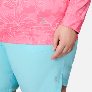 Wearing the Free Country Women's Plus Size SunFree UPF Hoodie in pink with a floral print and UPF 50+ sun protection, paired with light blue shorts, a person stands with their right hand resting near the pocket.
