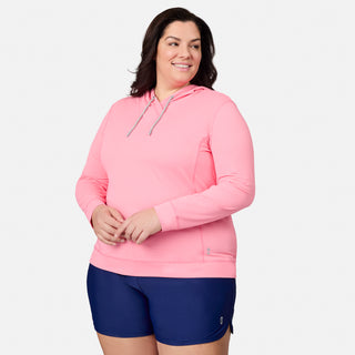 A woman wearing the Free Country Women's Plus Size SunFree UPF Hoodie in pink and navy shorts stands smiling against a plain white background, showcasing activewear designed with moisture-wicking and anti-odor properties.
