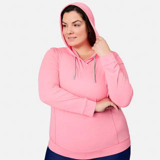 A woman wearing the Free Country Women's Plus Size SunFree UPF Hoodie stands against a plain white background, looking slightly to the side with one hand touching her hood. This light pink hooded sweatshirt features moisture wicking technology for all-day comfort and freshness.