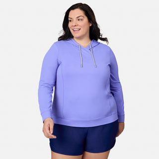 A woman is smiling against a plain white background, wearing the Free Country Women's Plus Size SunFree UPF Hoodie in light purple and dark navy shorts. The hoodie offers UPF 50+ sun protection and moisture-wicking technology to keep her cool and comfortable all day long.