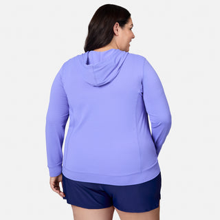 A woman is shown from the back, standing against a plain white background, wearing the Free Country Women's Plus Size SunFree UPF Hoodie in light purple and navy shorts. The hoodie features moisture-wicking properties to help keep her comfortable and dry throughout the day.