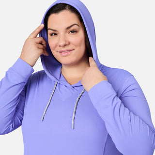 A woman smiles against a plain white background, wearing the Free Country Women's Plus Size SunFree UPF Hoodie in light purple, which features UPF 50+ sun protection. She holds the edge of her hood with one hand and touches her head with the other.