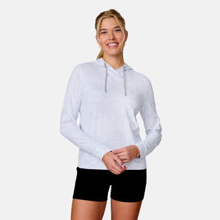 A smiling woman wears the Free Country Women's SunFree UPF Hoodie and black shorts, standing against a plain white background.
