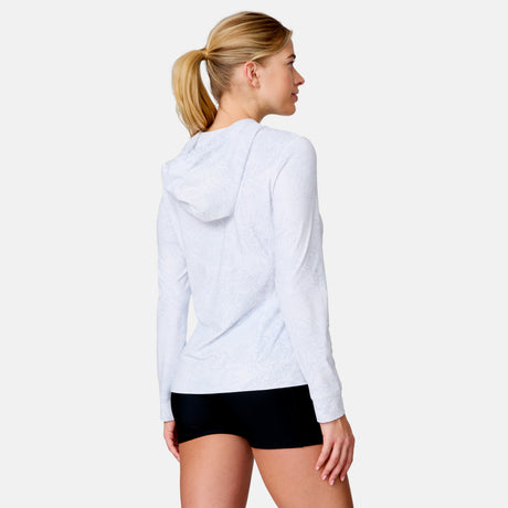 A woman with blonde hair in a ponytail wears the Free Country Women's SunFree UPF Hoodie in white and black shorts, standing with her back to the camera against a plain light background.