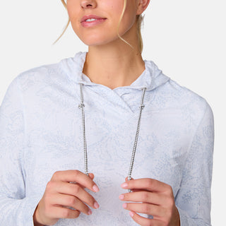A woman wearing the Free Country Women's SunFree UPF Hoodie in light blue holds both drawstrings and smiles gently. Her face is partly visible against a plain white background.