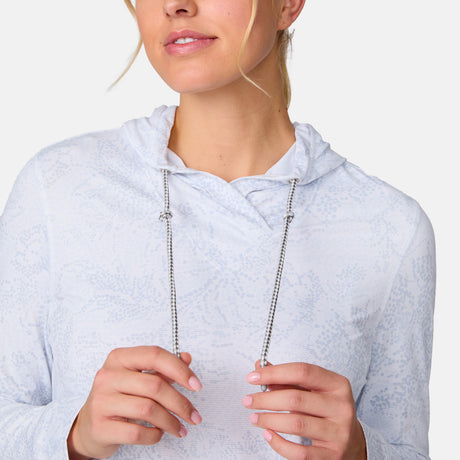 A woman wearing the Free Country Women's SunFree UPF Hoodie in light blue holds both drawstrings and smiles gently. Her face is partly visible against a plain white background.