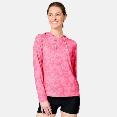A smiling woman wears the Free Country Women's SunFree UPF Hoodie, featuring long sleeves, a pink floral pattern, moisture-wicking fabric, and a drawstring, paired with black shorts against a light gray background.