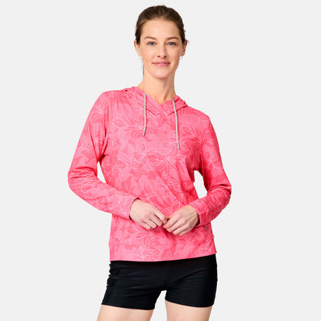 A woman smiles with her hands clasped, wearing the Free Country Women's SunFree UPF Hoodie in pink with subtle floral pattern and black shorts, standing against a plain light background.